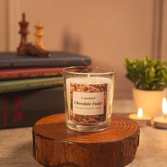 A 'Chocolate Fudge' scented candle by Radiate candles placed on a wooden stand with books, chess pieces, lit tealights and a plant in a warm cozy setting.