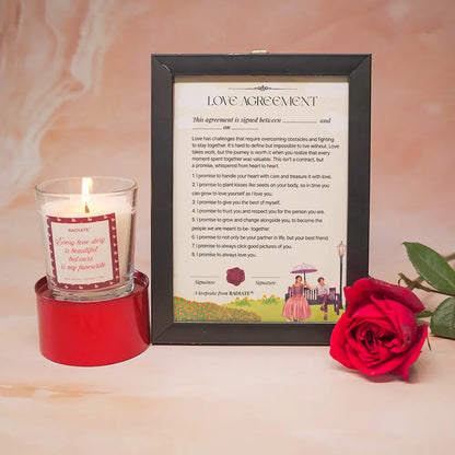 Framed 'Love Agreement' document with a lit love story scented candle and red rose on a soft pink background