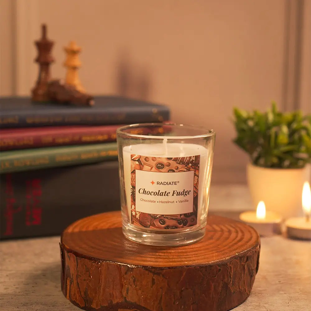 A 'Chocolate Fudge' scented candle by Radiate candles placed  on a wooden stand with books, chess pieces, lit tealights and a plant in a warm cozy setting. 