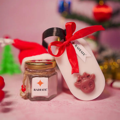 Radiate's Christmas essential duo with chocolate fudge scented candle jar wearing a Christmas hat and a Rudolf wax sachet with red ribbon. surrounded by Christmas decorations in background. 