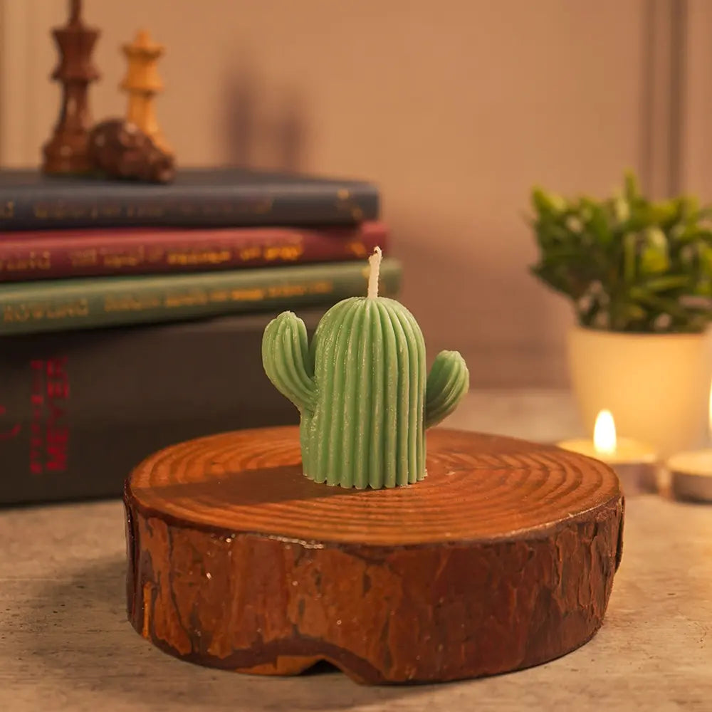 A Dancing Cactus-shaped designer pillar candle by Radiate on a wooden coaster with books and a plant in the background, warm cozy vibes. 