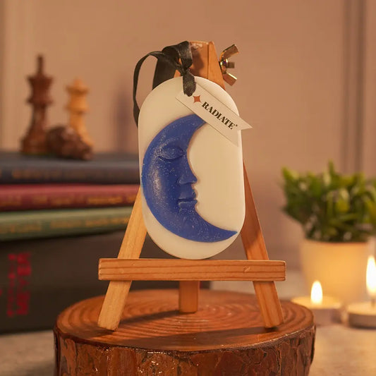Decorative moon face design embed on a scented wax sachet by Radiate on a wooden stand with a blurred background. Used for sweet scent in closed spaces like almirah, boxes, clothes, shoe rack, gym, tables etc