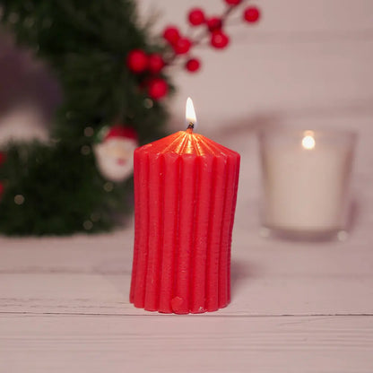 Red ribbed scented pillar candle by radiate  with a cotton wick on a wooden table, blurred Christmas decorations and lit candle in the background