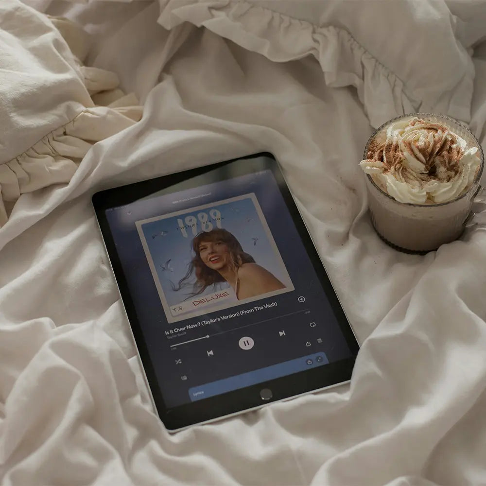 Tablet on a cozy bed displaying a music app with a cup of hot chocolate next to it.