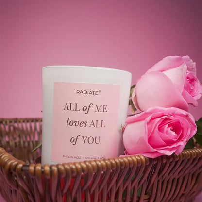 All of me scented candle in a basket surrounded by rose flowers on a pink background. 
