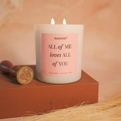 All of me lotus and musk scented candle with a pink label on a wooden block, with a soft peach background