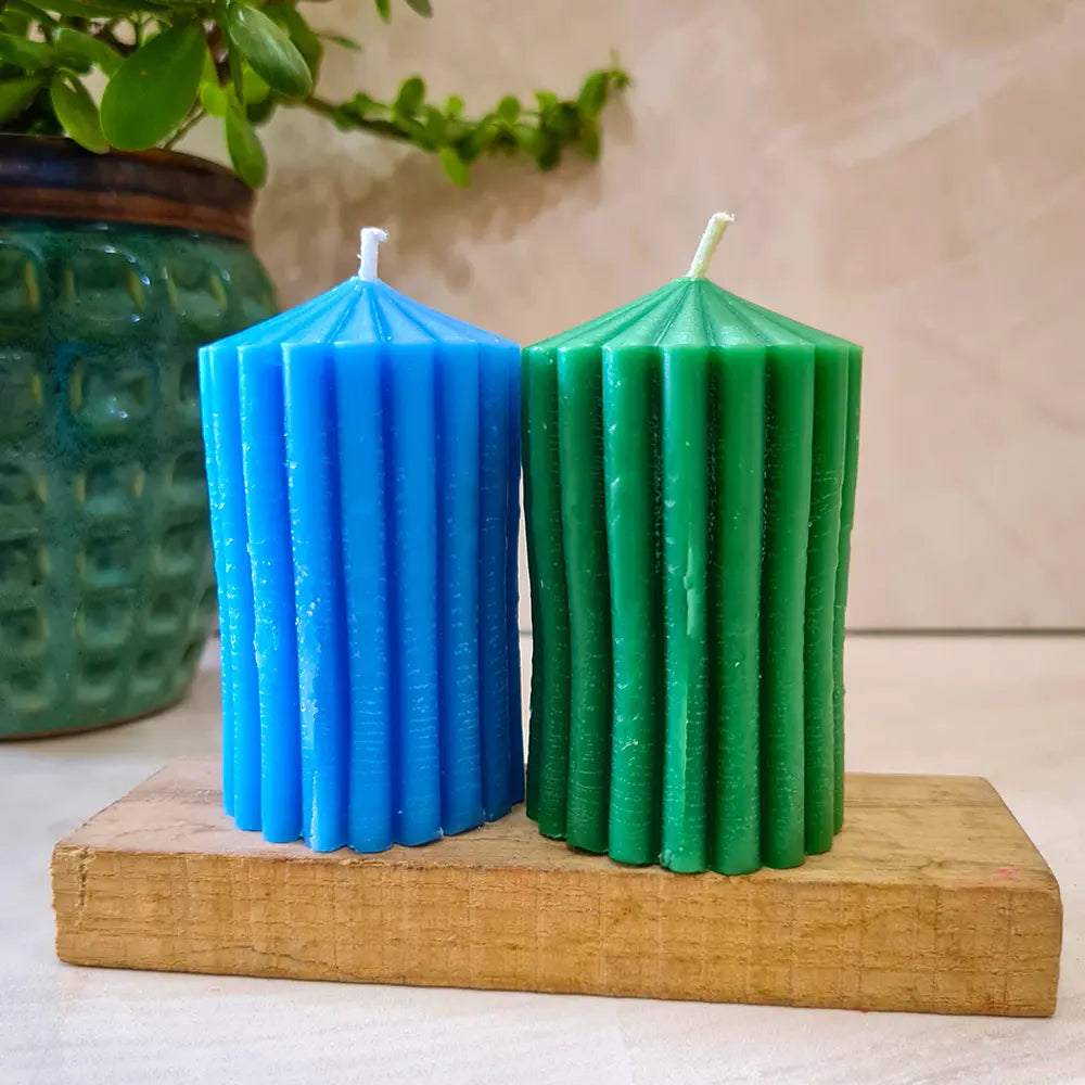 Two blue and green ribbed striped scented pillar candles on a wooden block with a plant in the background.
