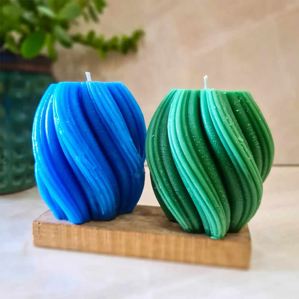 Two blue and green spiral swirl wave scented pillar candles on a wooden stand with a blurred plant in the background.