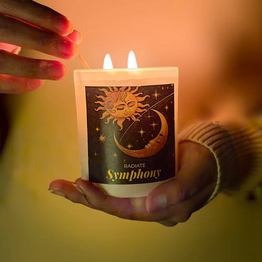 Radiate's Symphony-  Signature scented soy wax candle with aroma of Rose, Musk with celestial design labeled displaying union of sun and moon, held by a person with a warm glow lighting up the candle. 